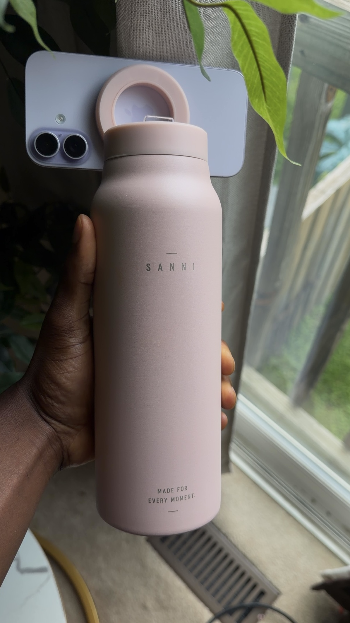 Pink SANNI bottle held beside a window with a phone attached