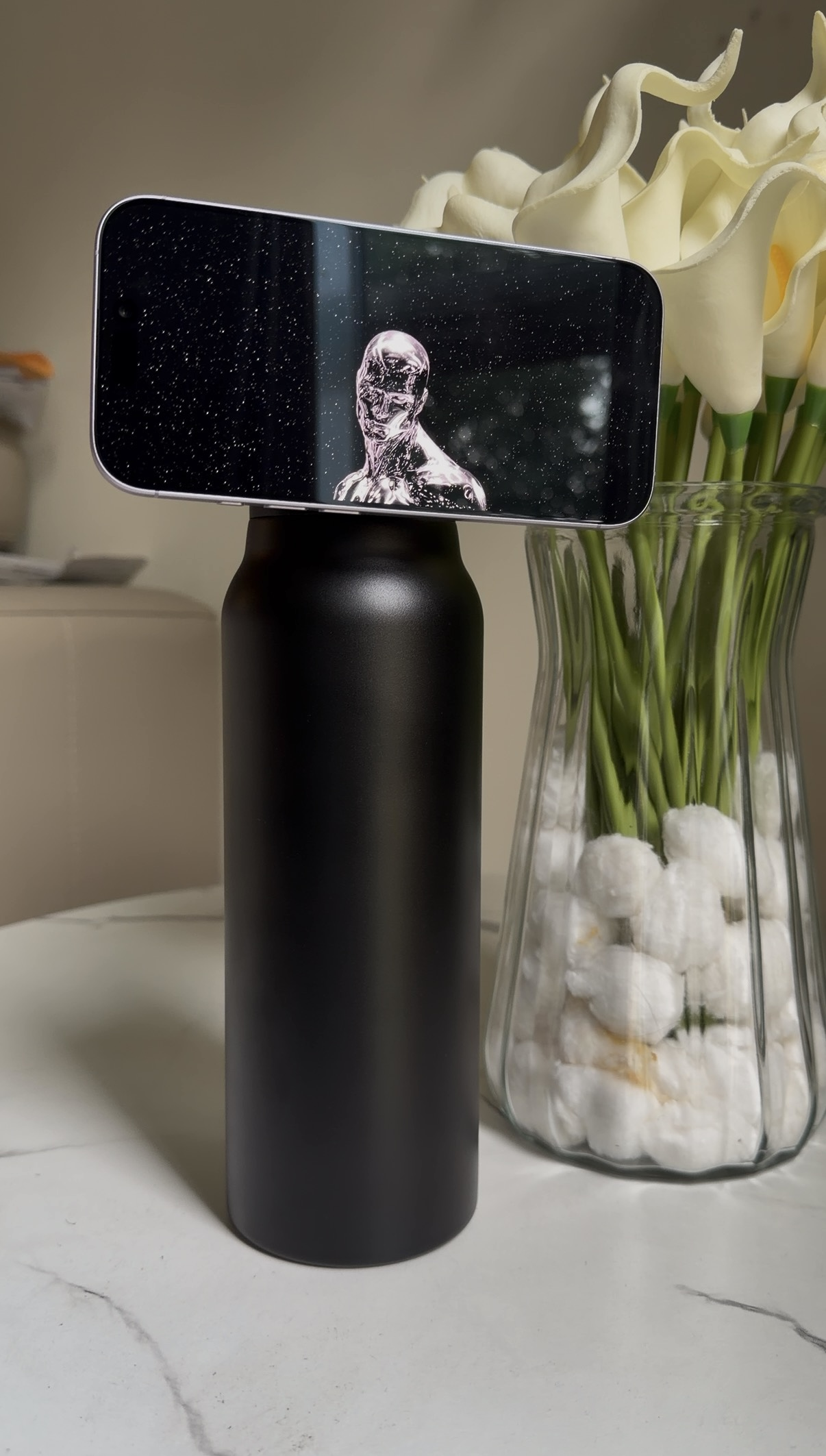 Black SANNI bottle supporting a phone beside white flowers