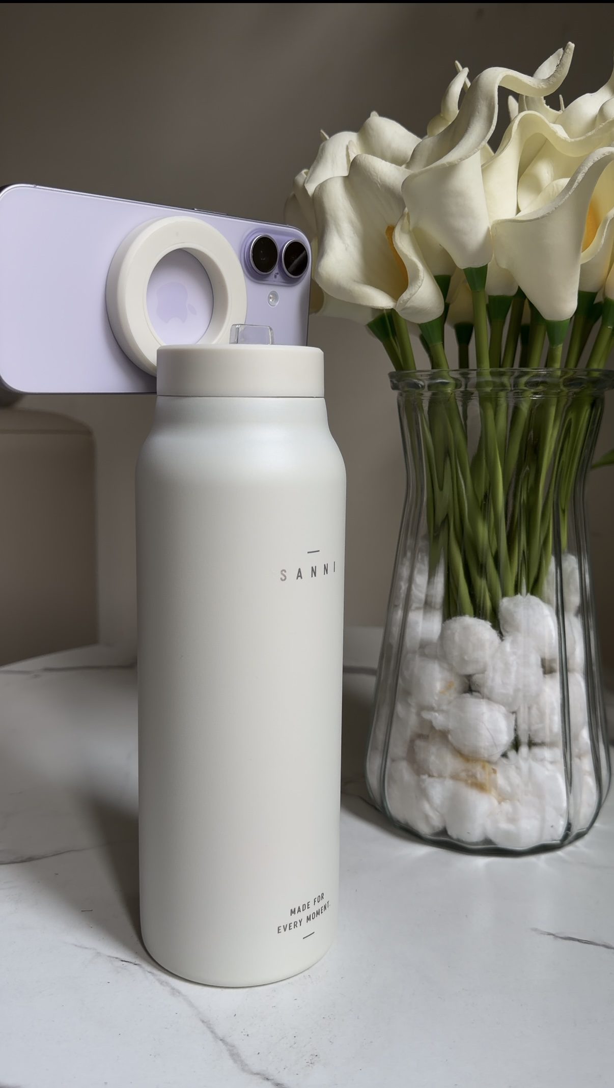 Cream White SANNI bottle magnetically holding a phone beside white flowers